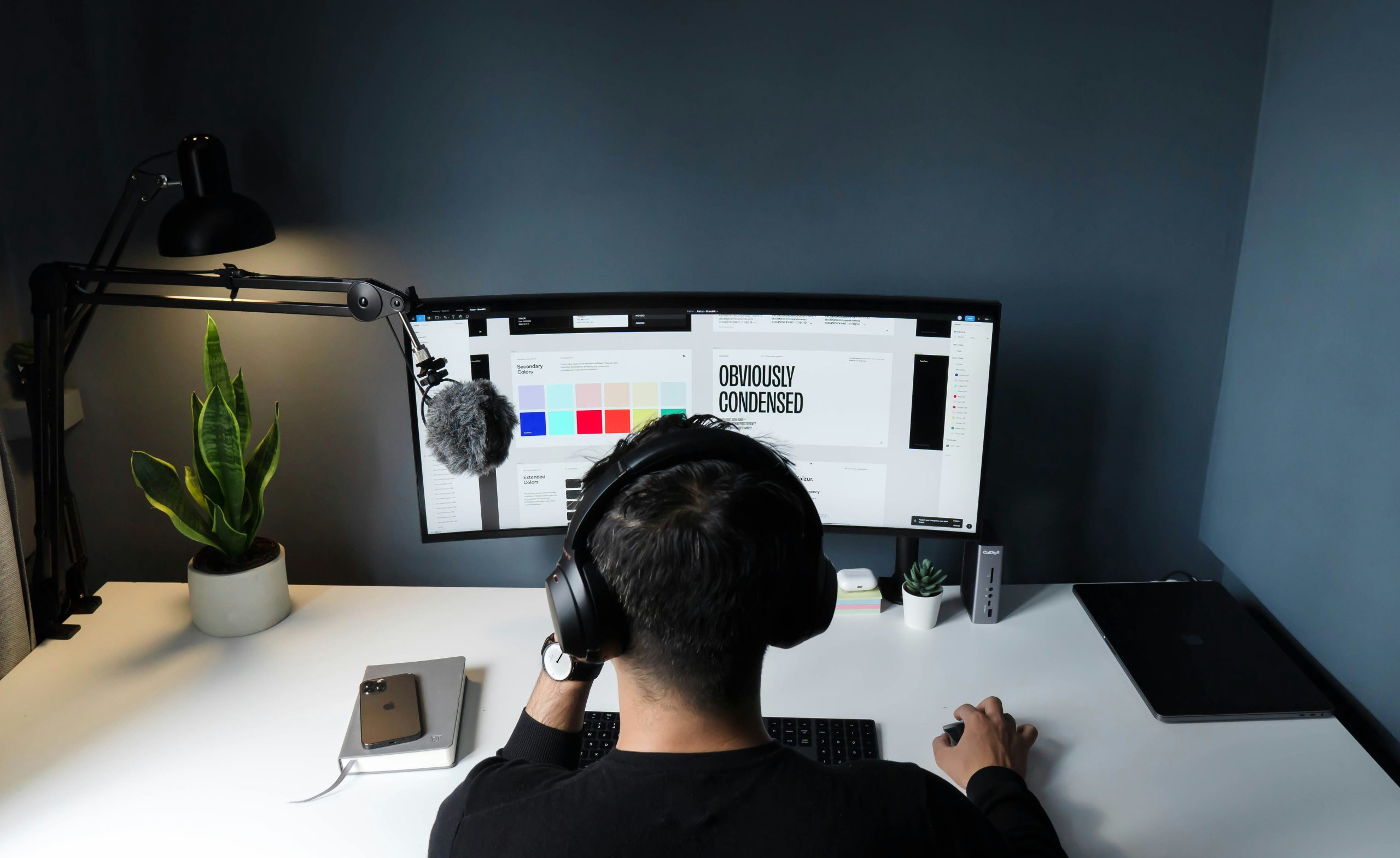 A freelancer working at a large ultrawide monitor in a dark home office setup with headphones on