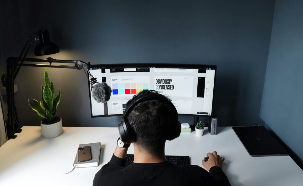 A freelancer working at a large ultrawide monitor in a dark home office setup with headphones on